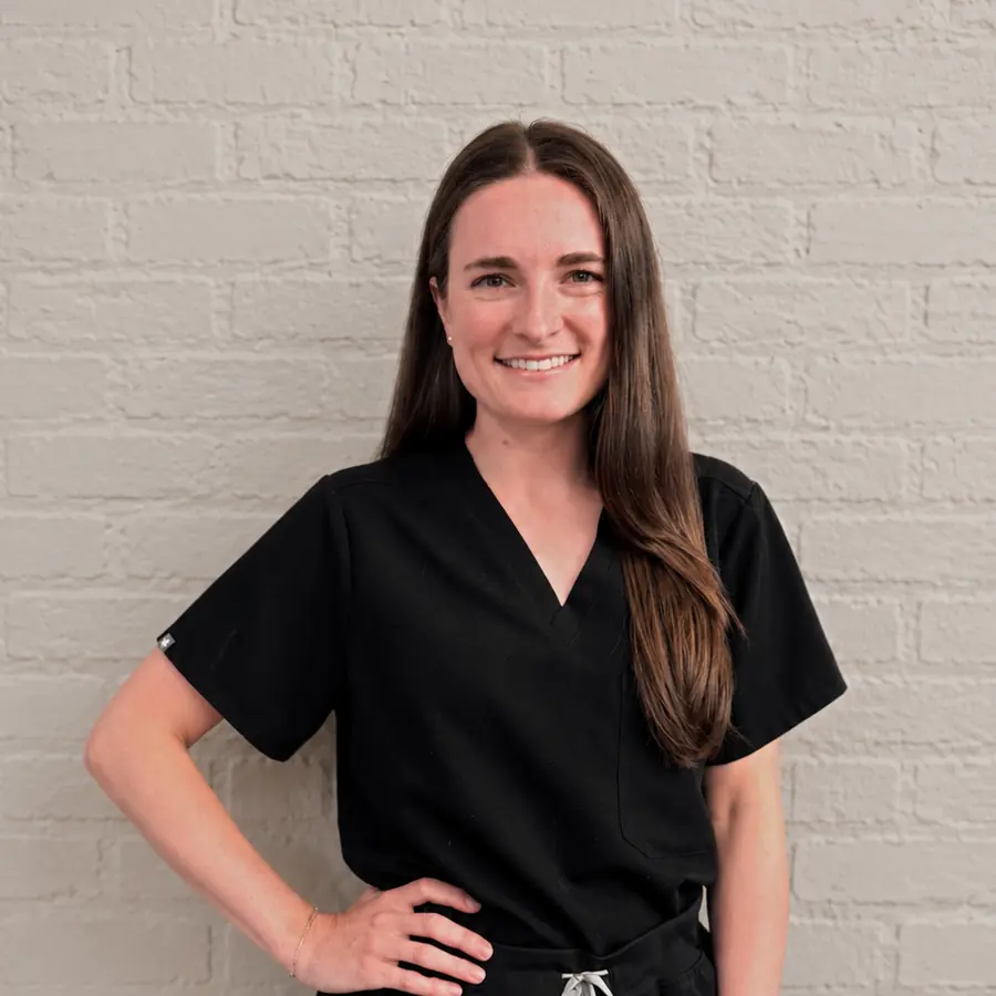 Smiling woman with long brown hair in black scrubs, standing with a hand on her hip against a light brick wall background.