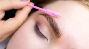 Close-up of a person with a pink brow brush shaping their eyebrow over a closed eye.