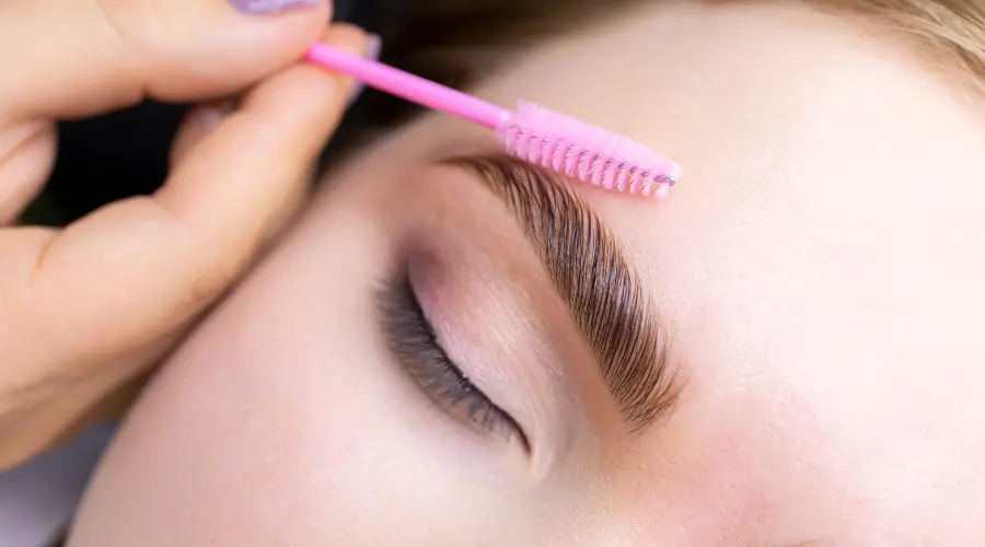 Close-up of a person with a pink brow brush shaping their eyebrow over a closed eye.