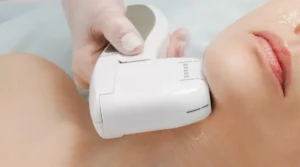 Close-up of a white handheld cosmetic device pressed against a woman's neck during a treatment, held by a practitioner